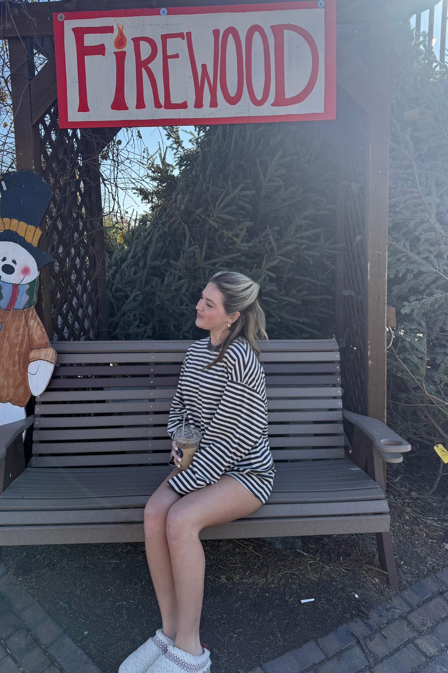 Woman in striped oversized sweater enjoying coffee on a bench near a firewood sign.