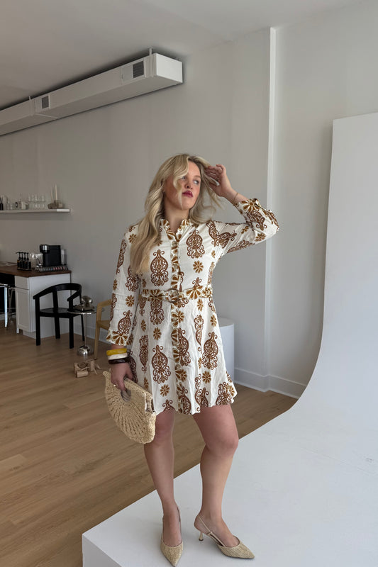Woman in a patterned dress standing in a room with wooden flooring and white walls.