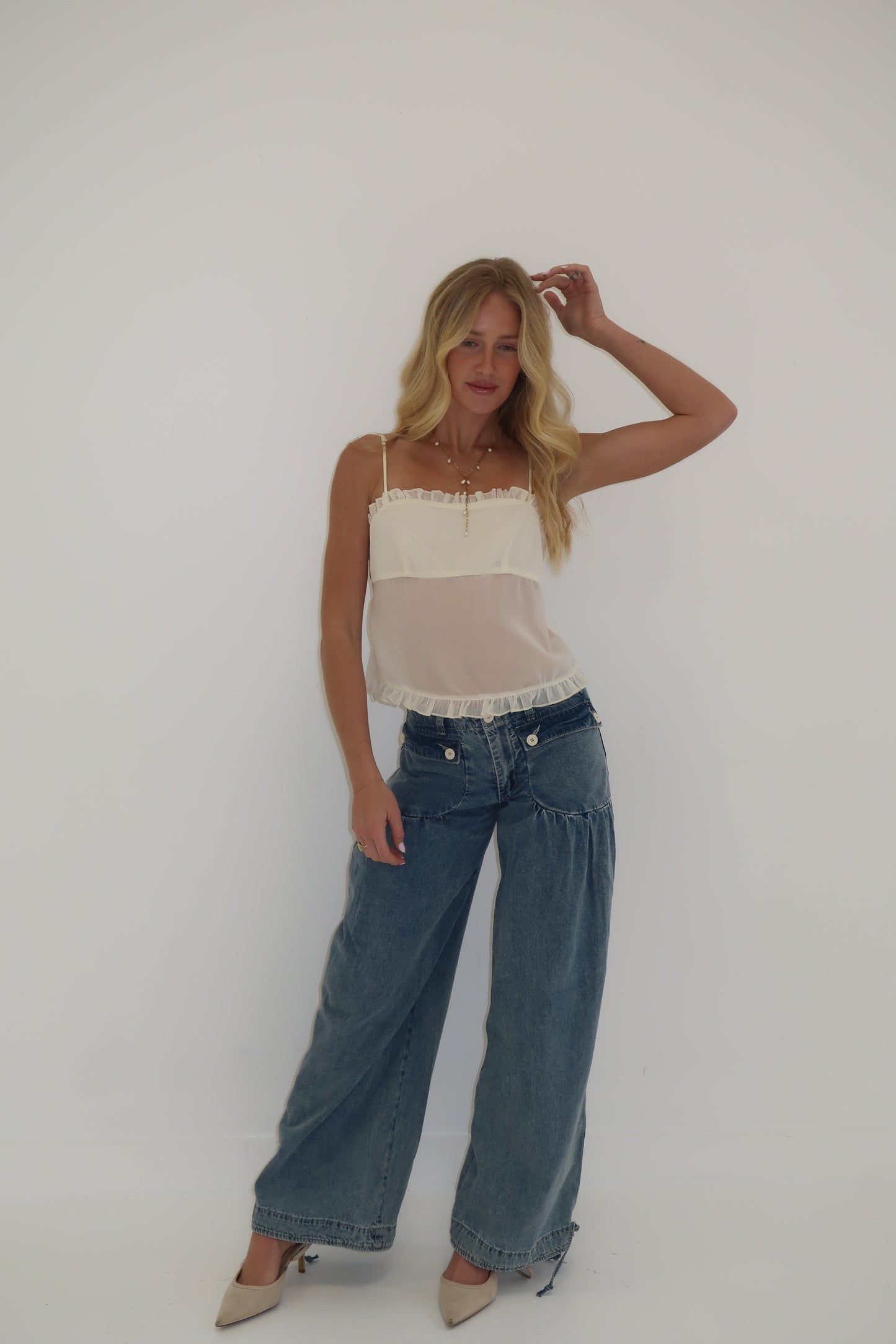 Model wears a cream sheer camisole with ruffle hem and wide-leg light blue jeans, styled with nude heels.