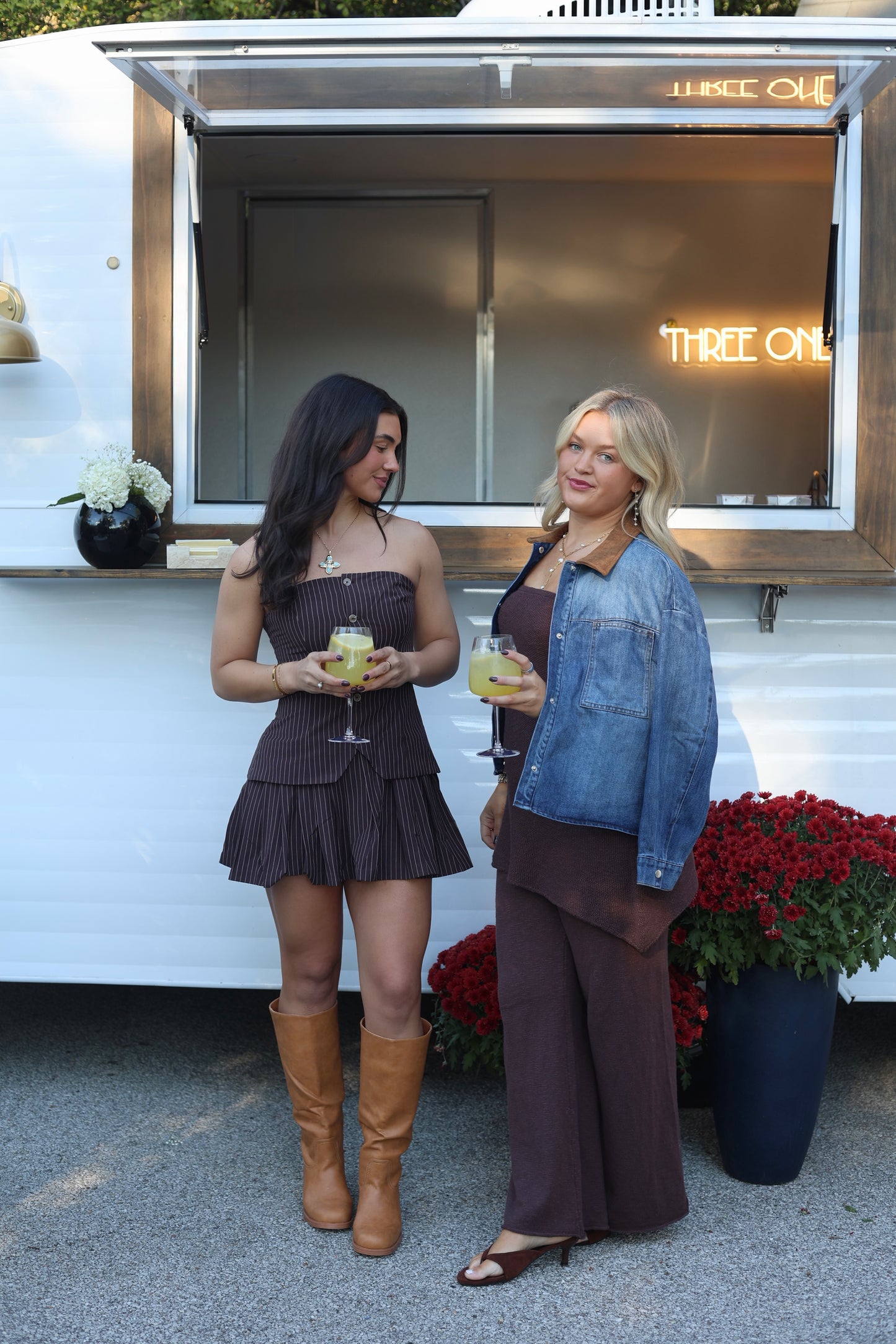 Two women enjoying drinks, one in a brown striped dress and the other in a denim jacket with brown pants.