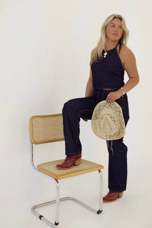 A woman wearing a dark denim halter jumpsuit and red western boots poses with one foot on a woven cane chair. She holds a straw cowboy hat in one hand and wears gold jewelry with a white cross pendant. Her blonde hair is styled down, and she gazes slightly off to the side against a plain light background. The overall look gives off a modern cowgirl vibe.