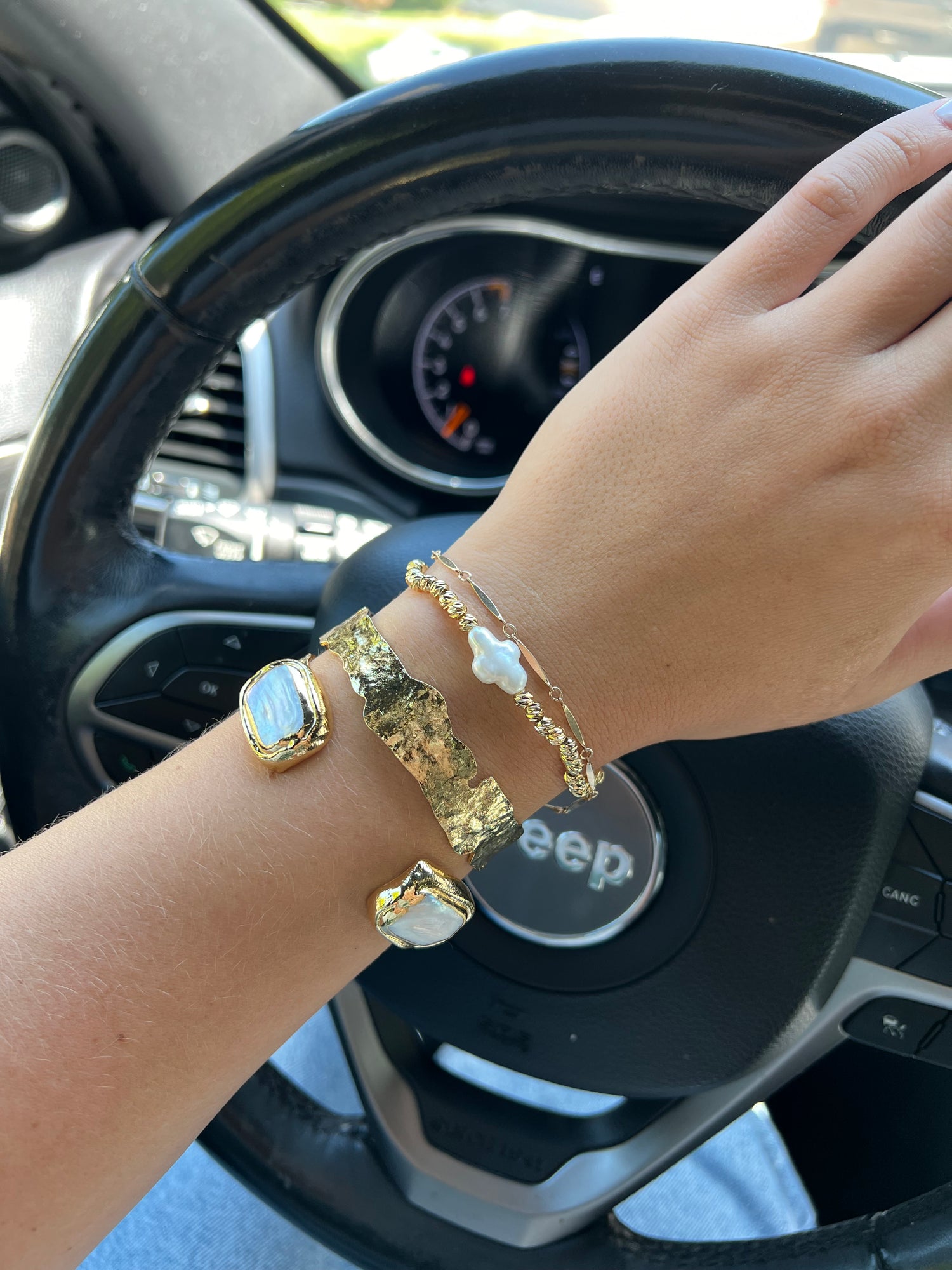 A close-up of a person's wrist wearing three bracelets: a chunky gold cuff with white stones, a delicate gold chain with a fresh water pearl cross charm, and a simple gold beaded