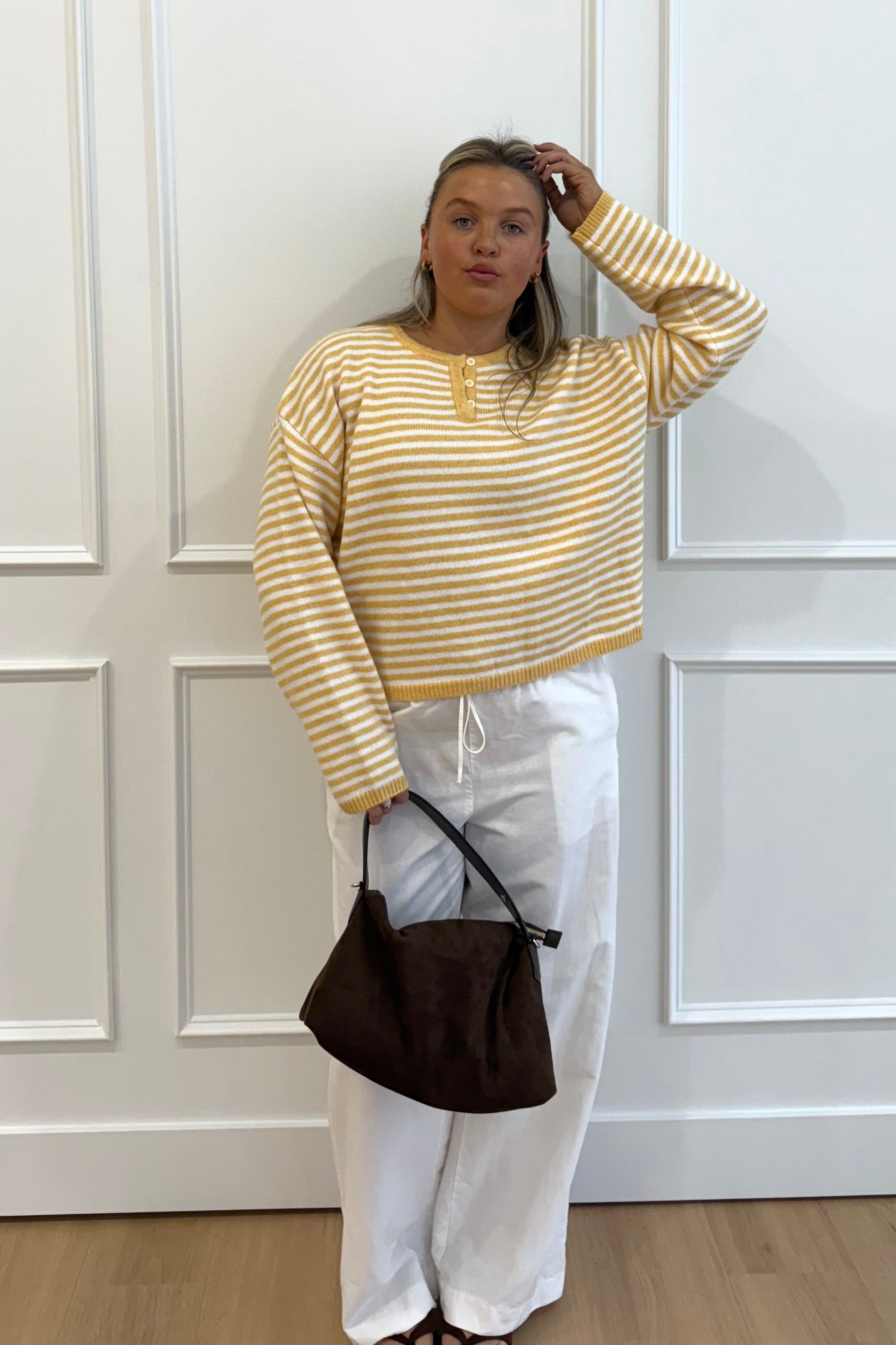 Yellow and white striped cropped sweater with buttons, paired with white pants and a brown handbag.