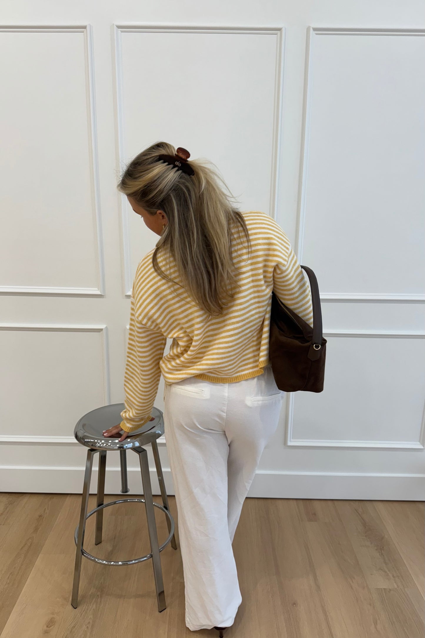 Woman wearing a yellow striped sweater and white pants, standing next to a chrome stool.