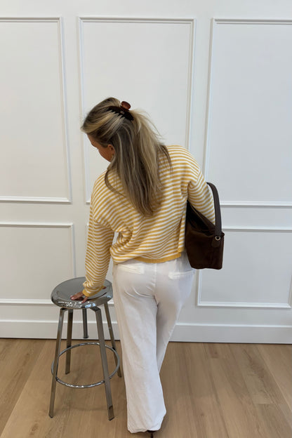 Woman wearing a yellow striped sweater and white pants, standing next to a chrome stool.