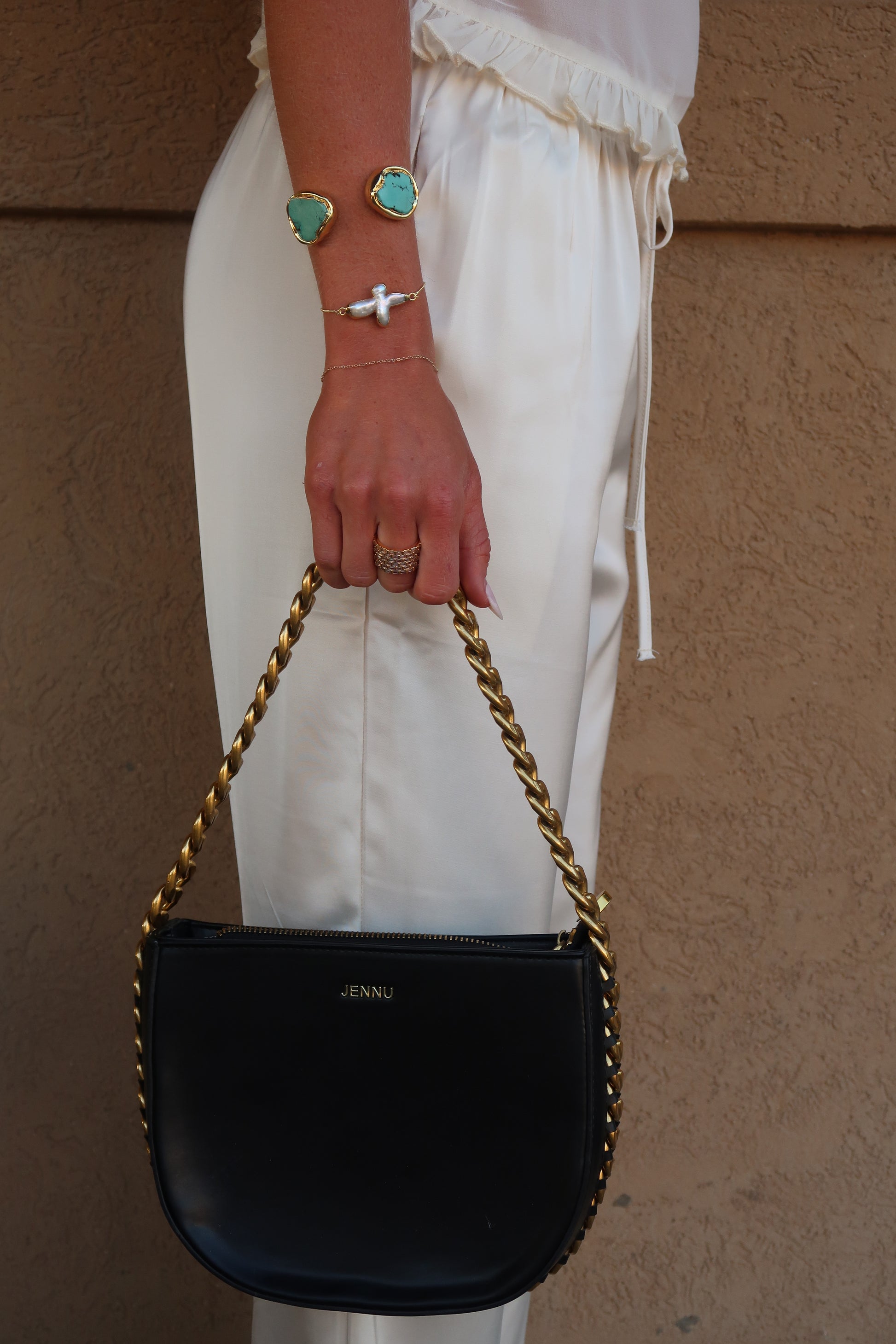 Black handbag with gold chain held by a hand wearing a ring and bracelet on a neutral background