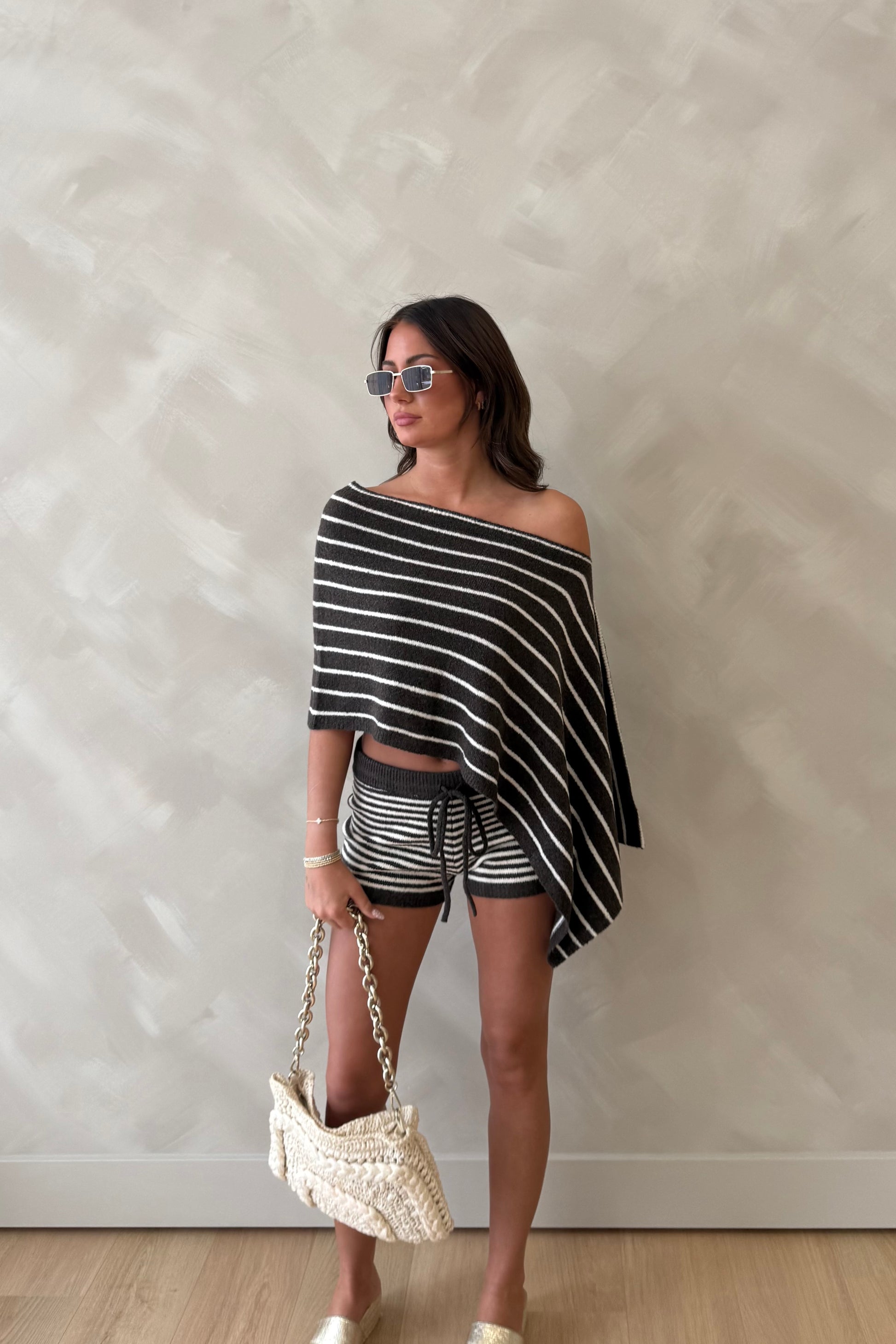 Woman wearing a black and white striped poncho with shorts and holding a handbag against a plain wall.
