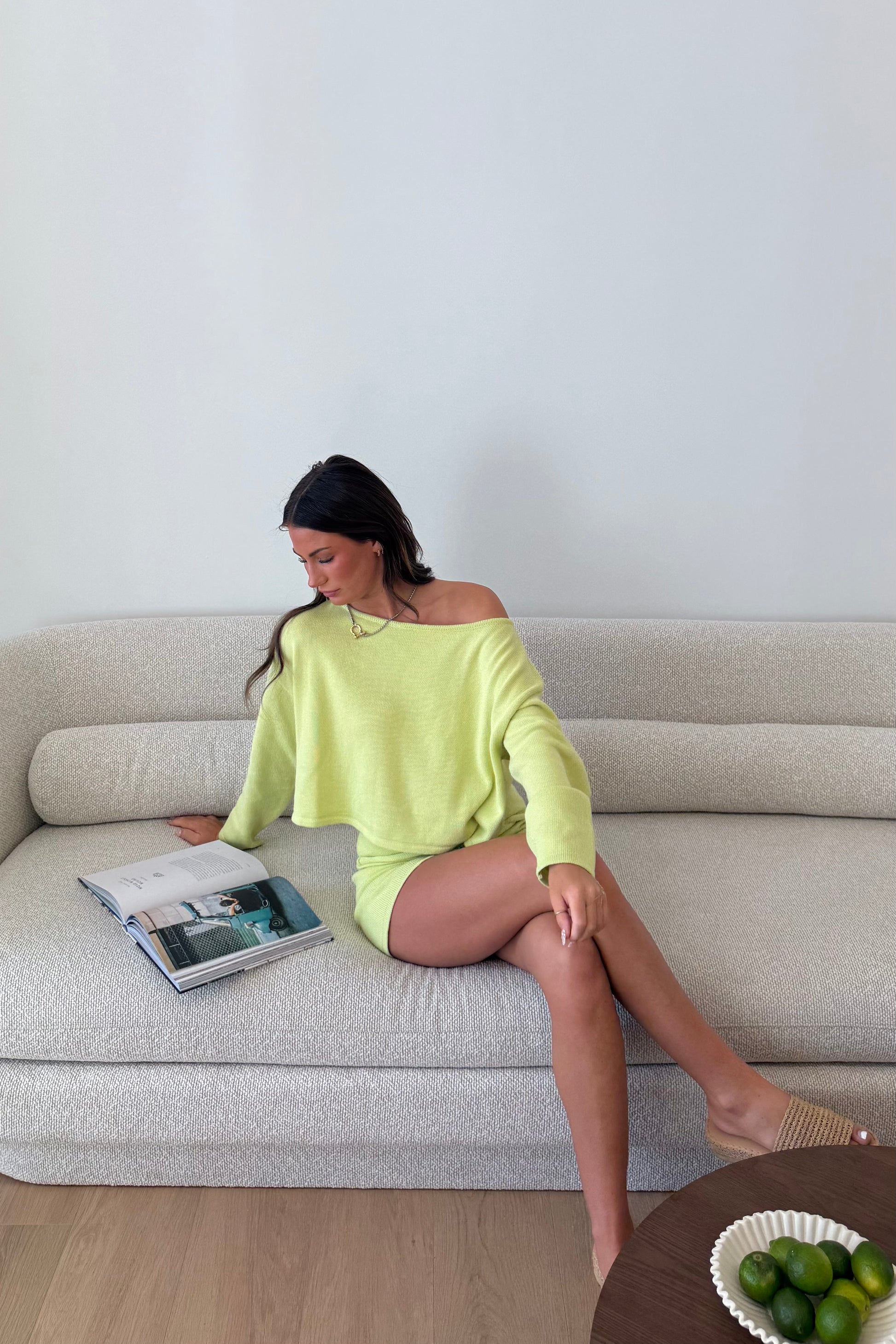 Woman in a bright green dress sitting on a beige sofa with a magazine, in a room with a light gray wall.
