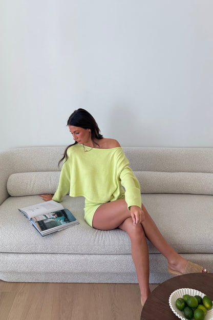 Woman in a bright green dress sitting on a beige sofa with a magazine, in a room with a light gray wall.