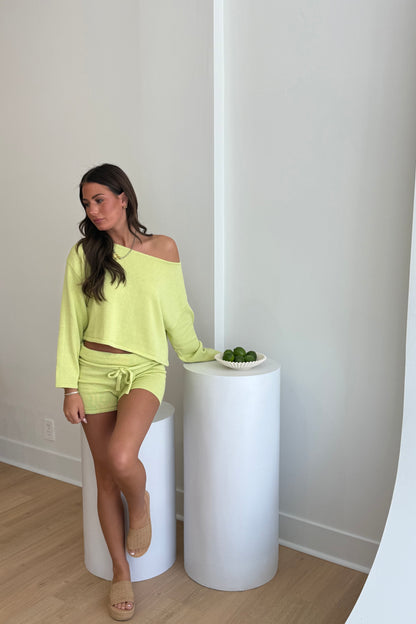 Woman in a bright yellow outfit standing next to a white pedestal with a bowl of fruit on a light wood floor.