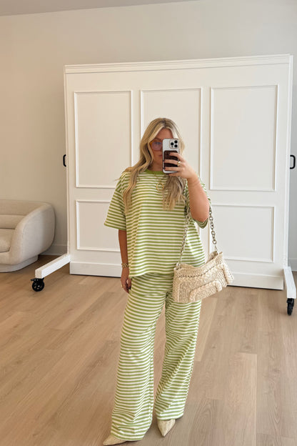 Woman taking a mirror selfie wearing a green and white striped outfit in a room with wooden flooring and white walls.