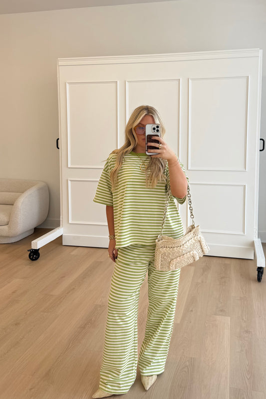 Woman taking a mirror selfie wearing a green and white striped outfit in a room with wooden flooring and white walls.