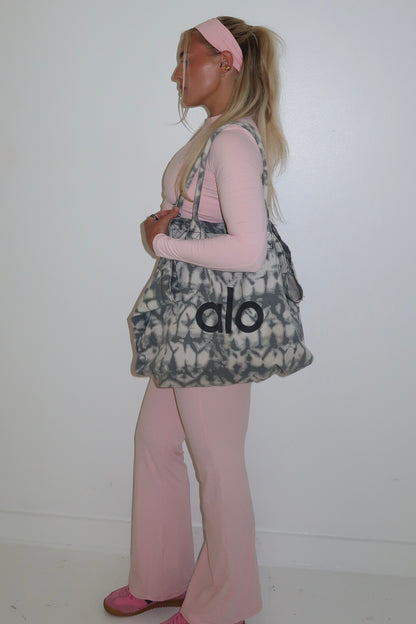 Person wearing a patterned bag with 'alo' logo, pink top, and pants on a white background