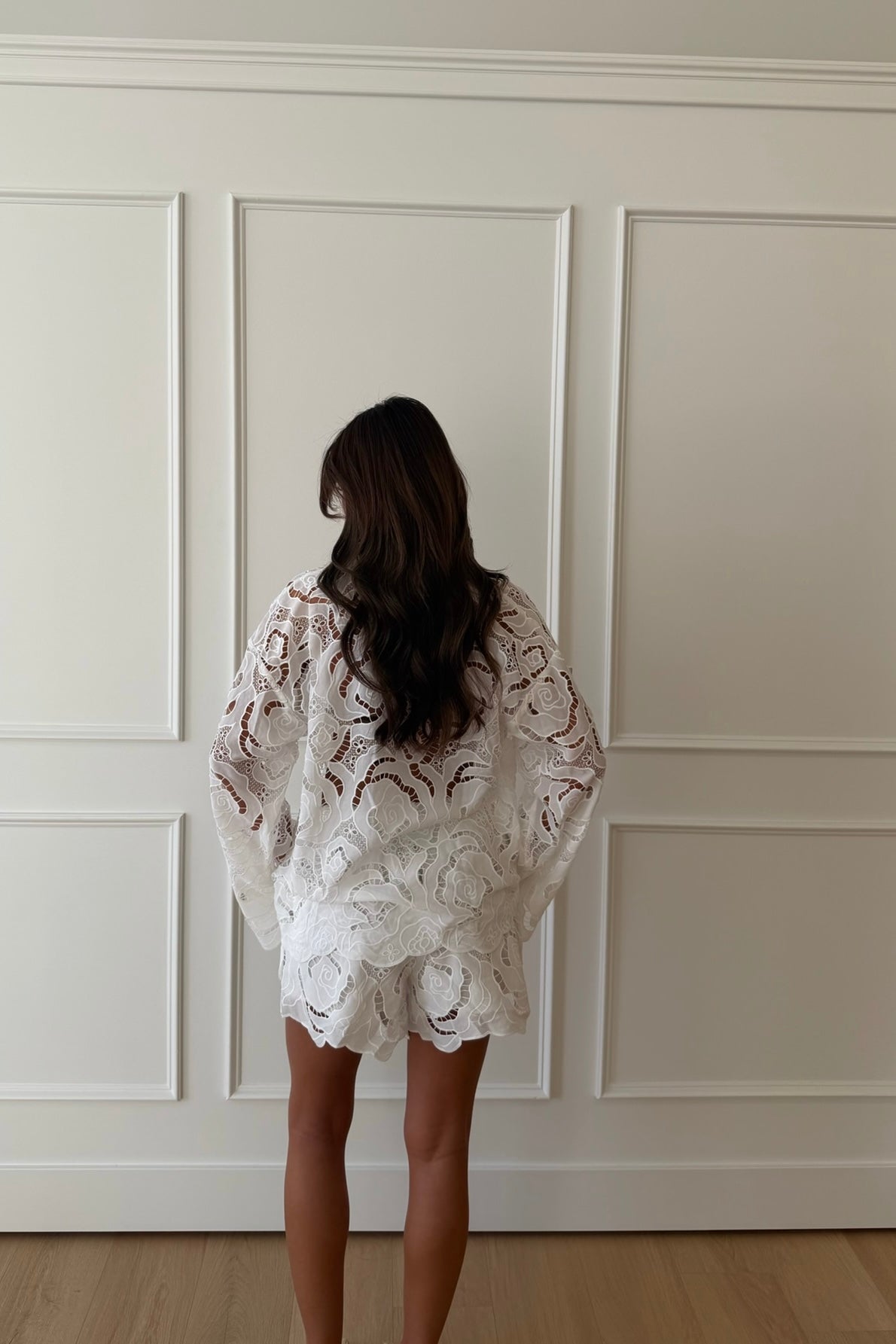 Woman wearing a white lace outfit standing against a paneled wall.