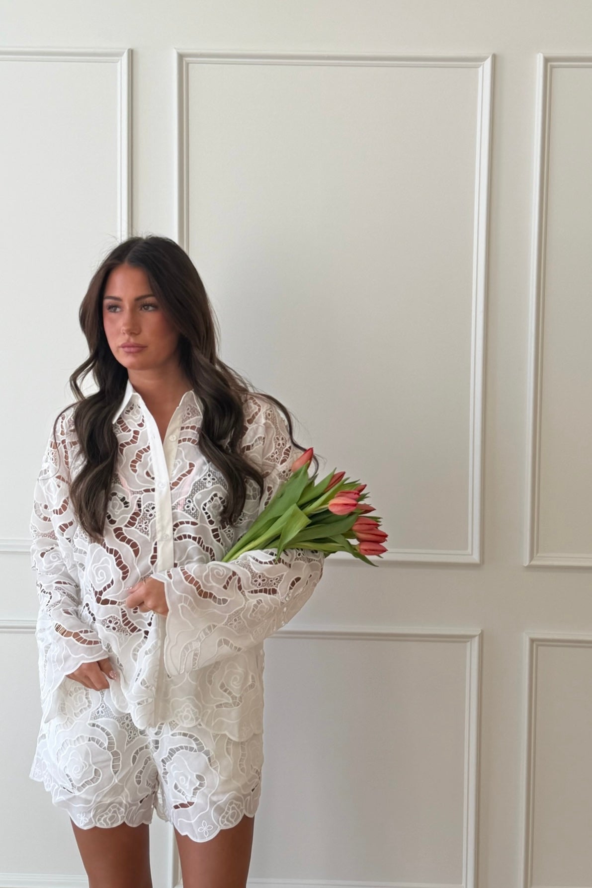 Woman in a white lace outfit holding red tulips against a white paneled wall.