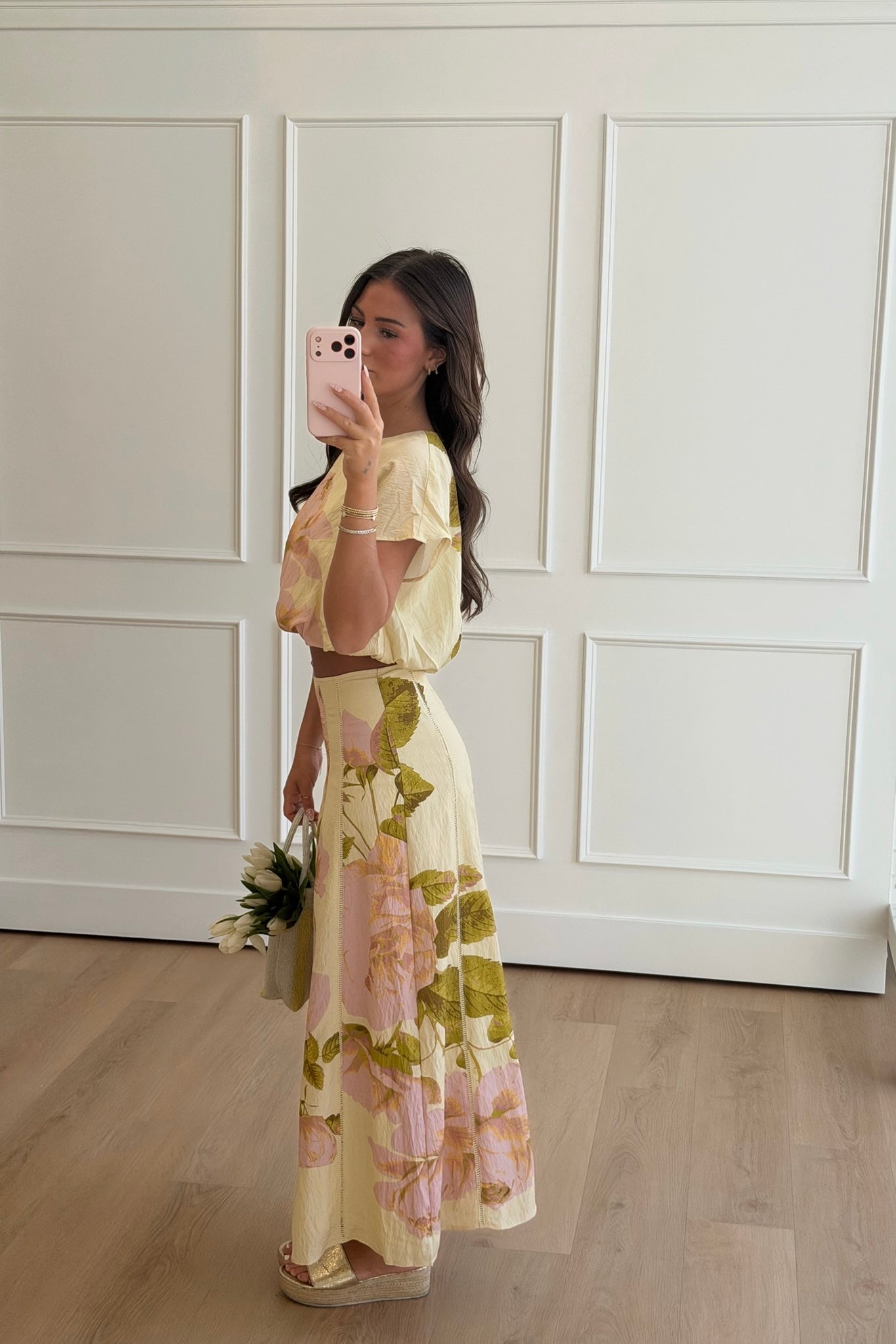 Woman taking a mirror selfie wearing a floral dress in a room with white paneled walls.