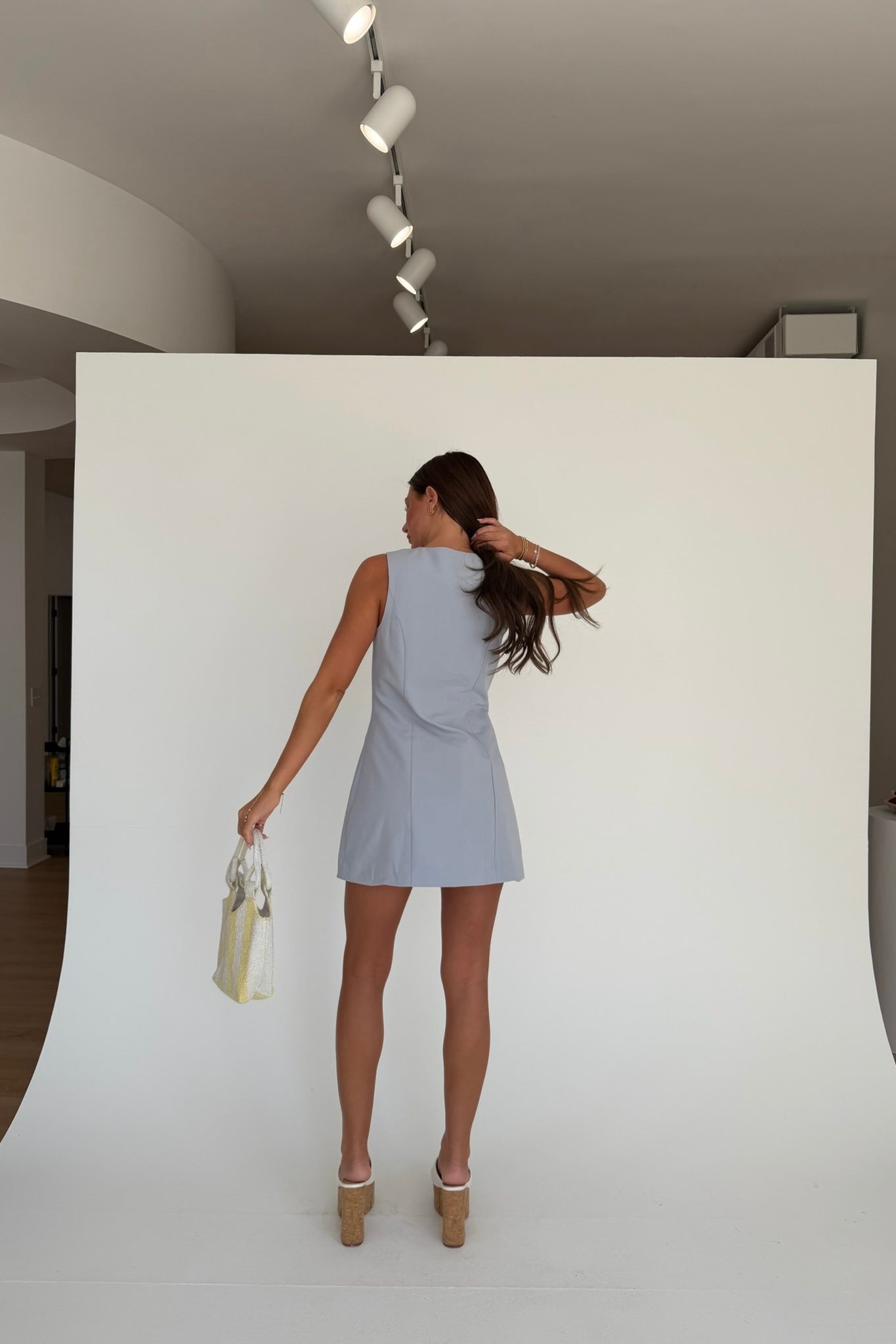 Woman in a light blue dress standing against a white backdrop in a minimalistic room.