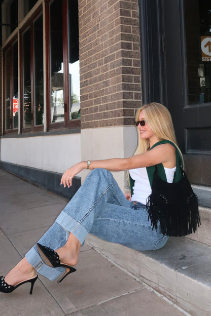 Woman sitting on a sidewalk wearing high heels, jeans, and a black fringed bag.