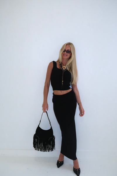 Stylish woman in black crop top and maxi skirt, holding a fringed black handbag, exuding chic elegance.
