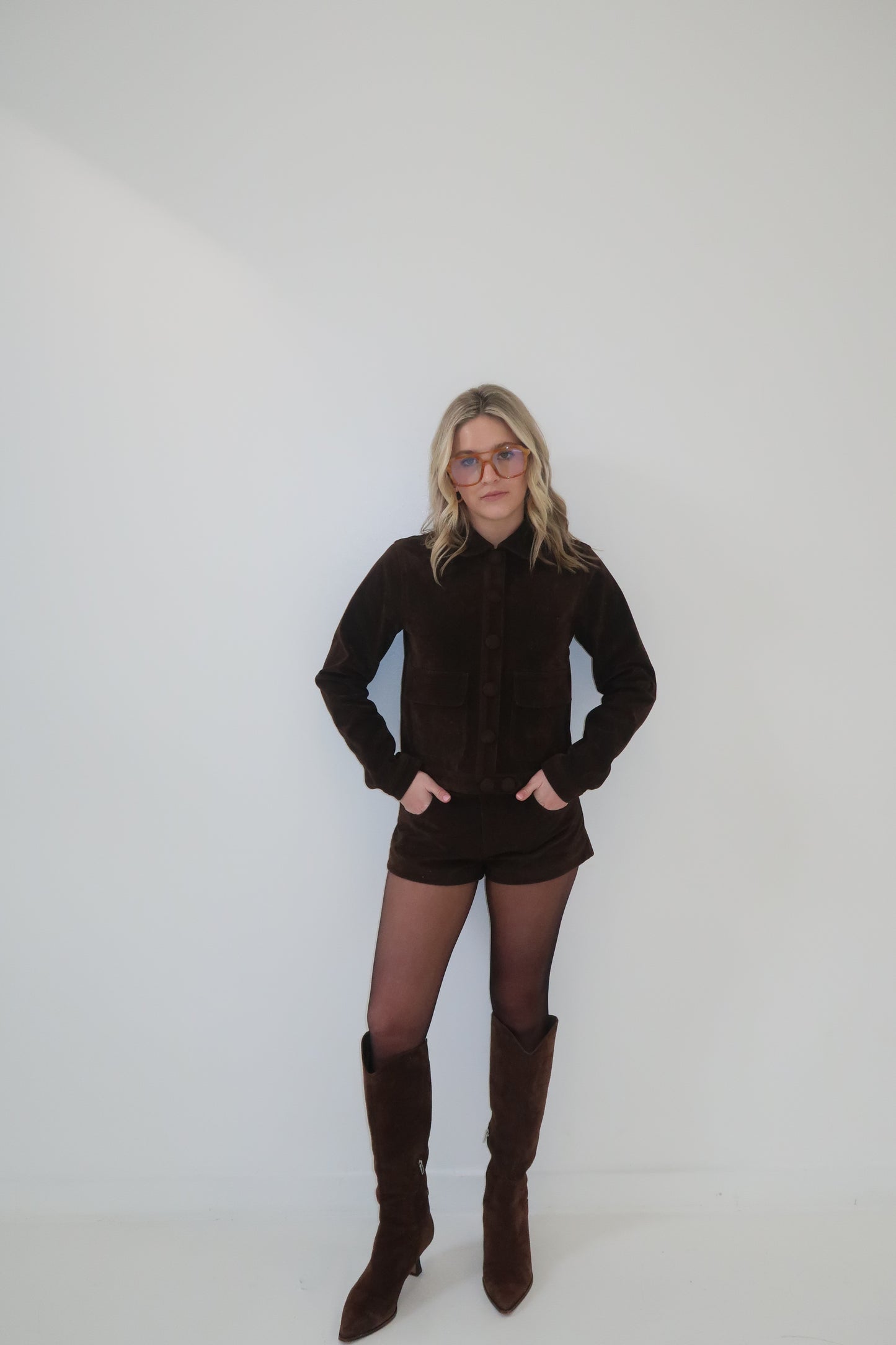Brown suede jacket and shorts set with button details, paired with knee-high boots and tinted glasses.