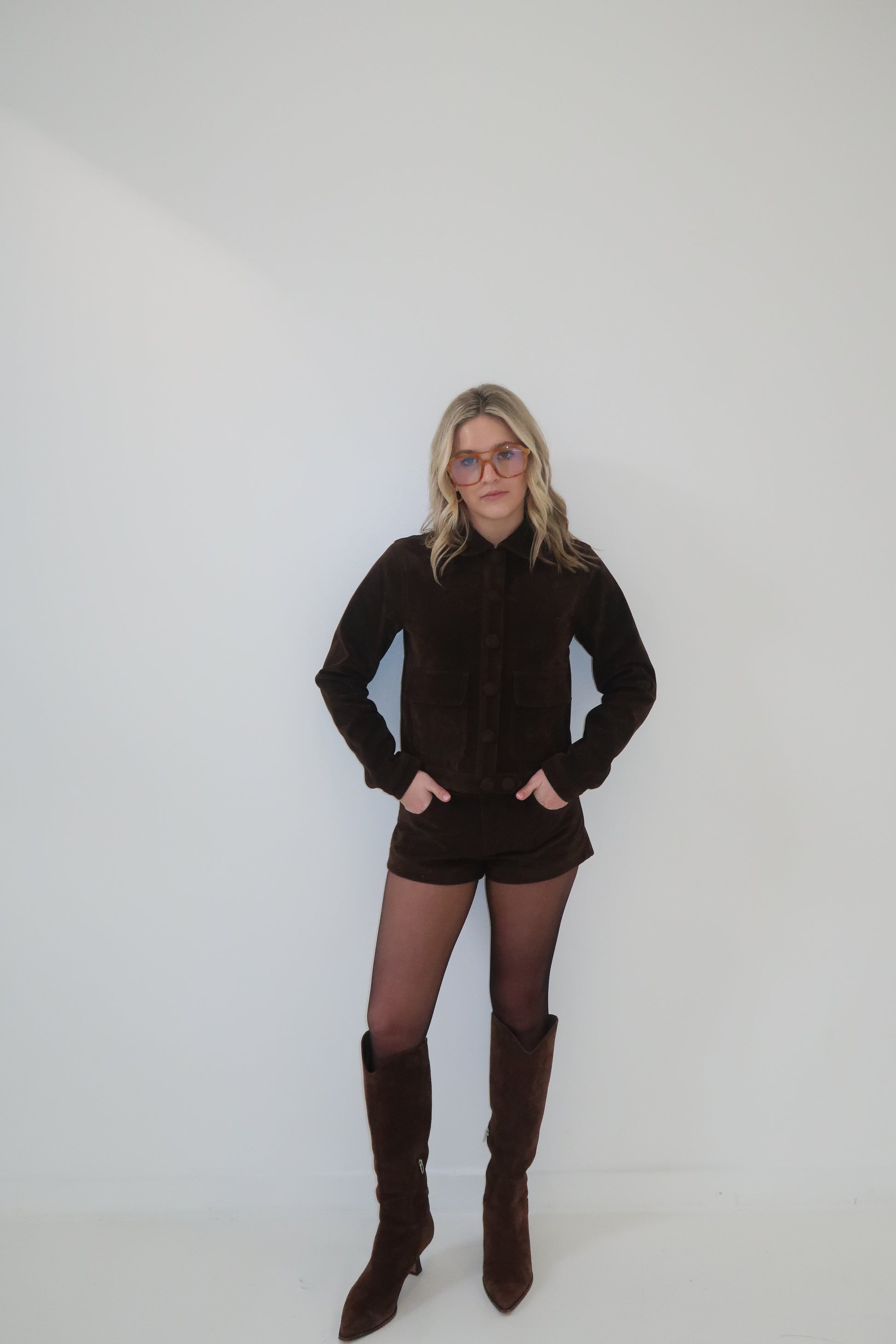 Brown suede jacket and shorts set with button details, paired with knee-high boots and tinted glasses.