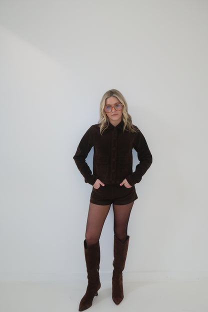 Brown suede jacket and shorts set with button details, paired with knee-high boots and tinted glasses.