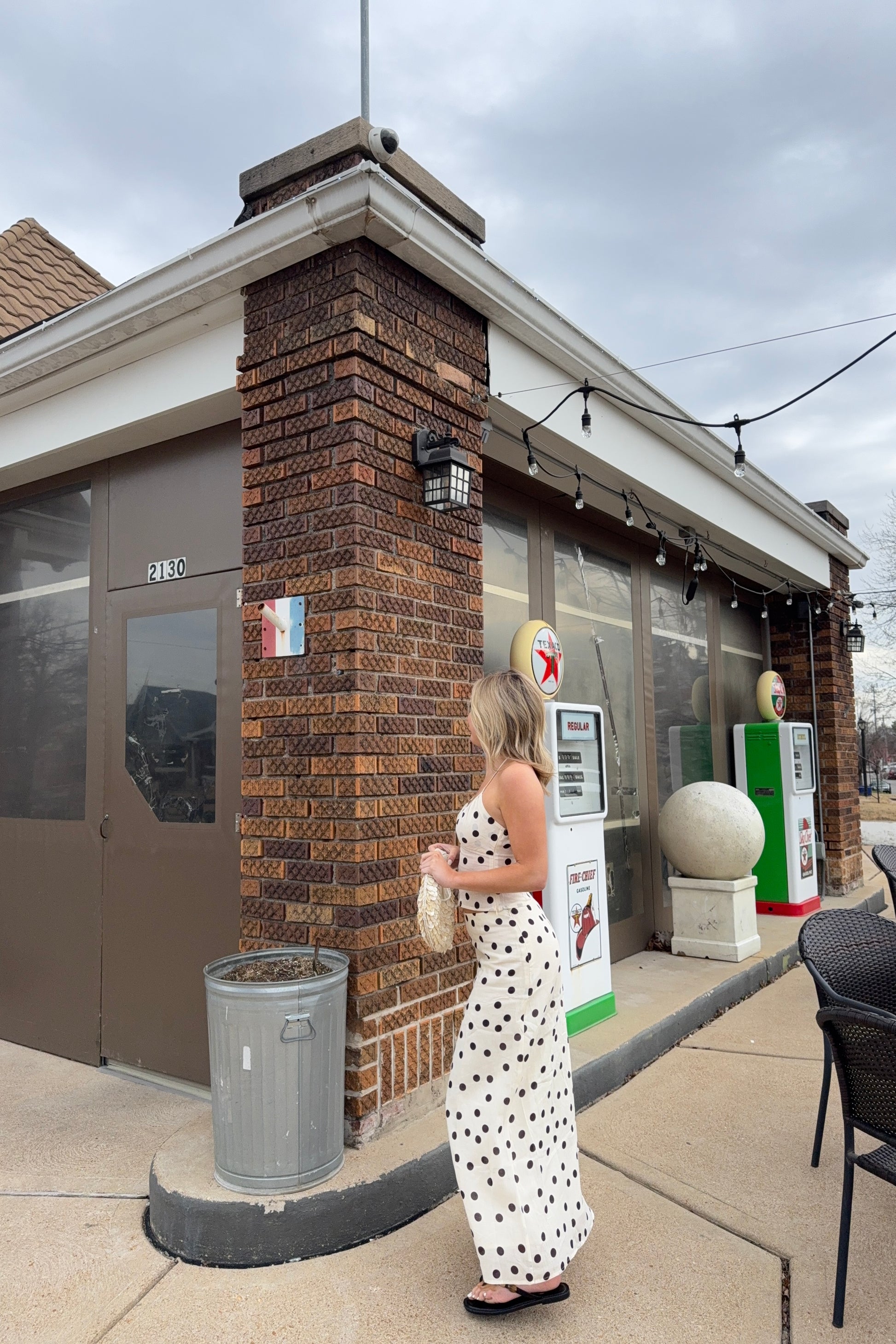 Woman in a polka dot dress standing outside a small building with a brick facade.