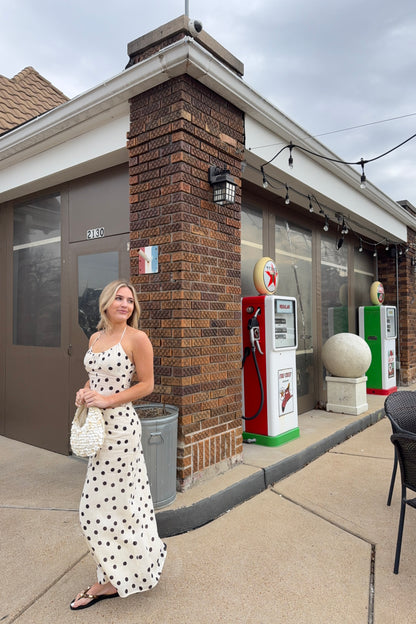 Woman in a polka dot dress standing outside a small building with vintage gas pumps.
