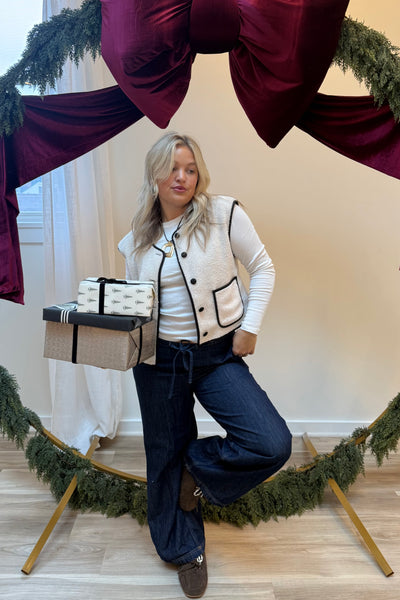 Woman wearing a cream vest, white long sleeve shirt, and dark denim pants, holding festive gift boxes.