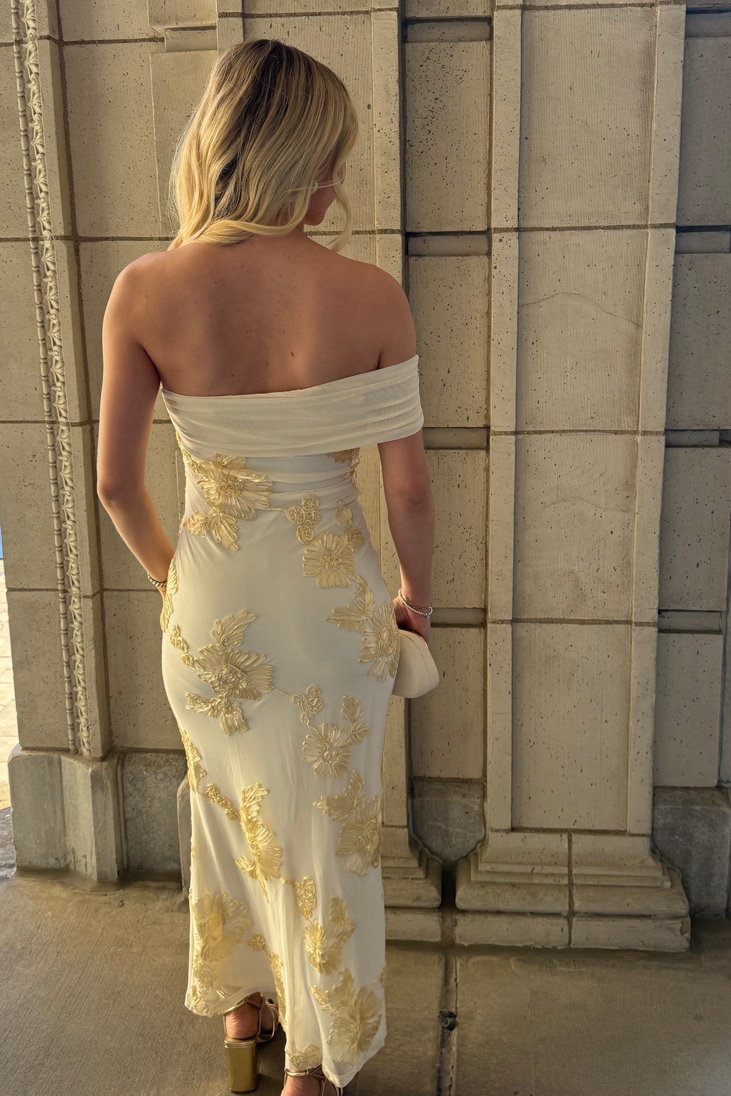 Woman in a white strapless dress with gold embroidery standing against a stone wall.