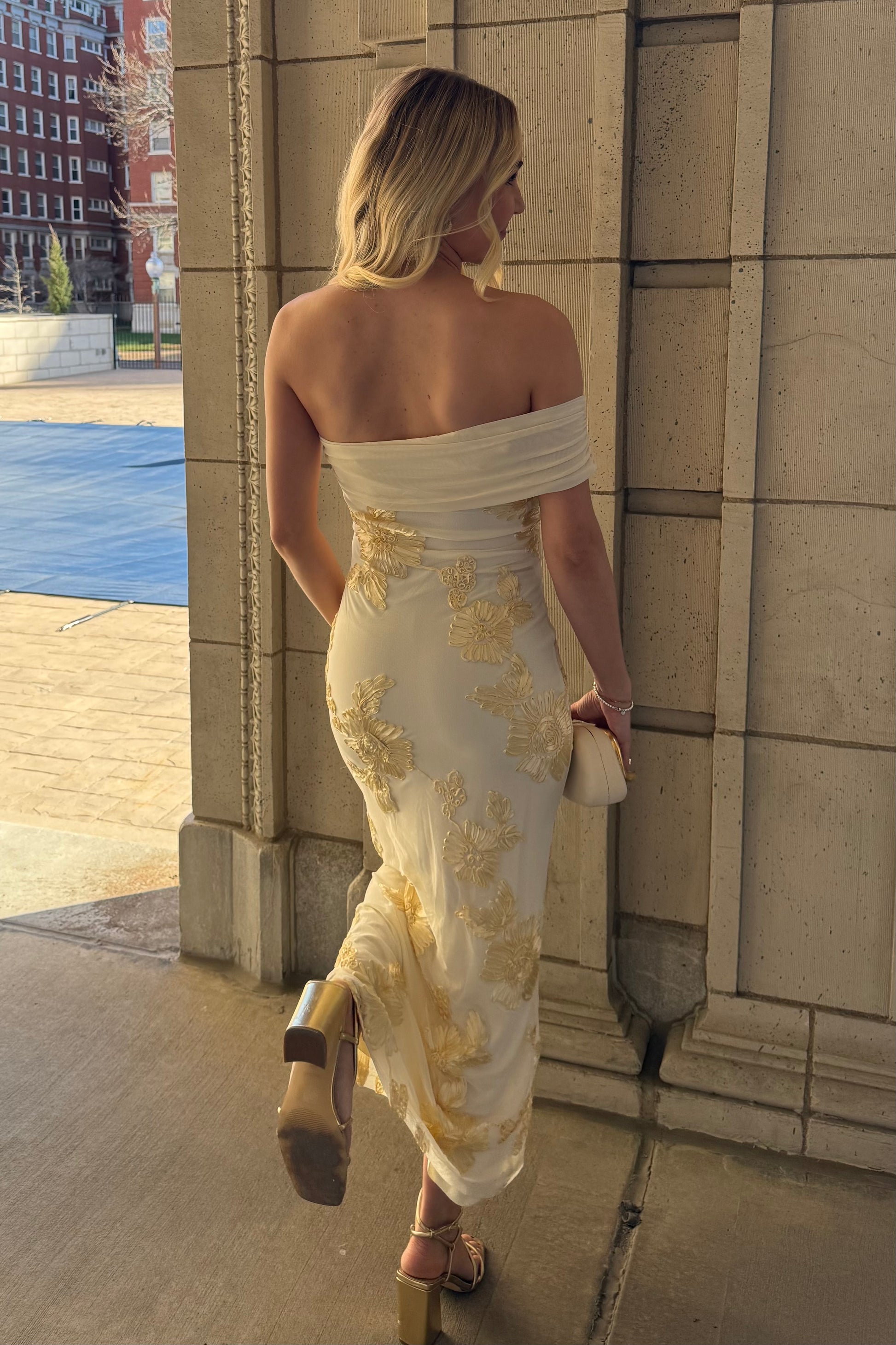 Woman in a white off-shoulder dress with gold embroidery standing against a building.