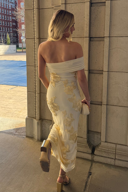 Woman in a white off-shoulder dress with gold embroidery standing against a building.