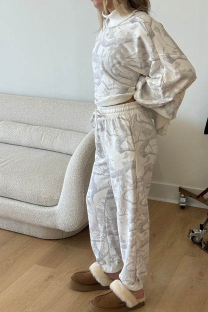 Person wearing a white patterned tracksuit standing in a room with a light-colored sofa.