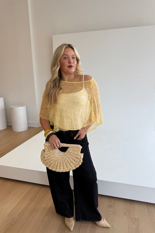 Woman wearing a yellow top and black pants, holding a woven handbag indoors.