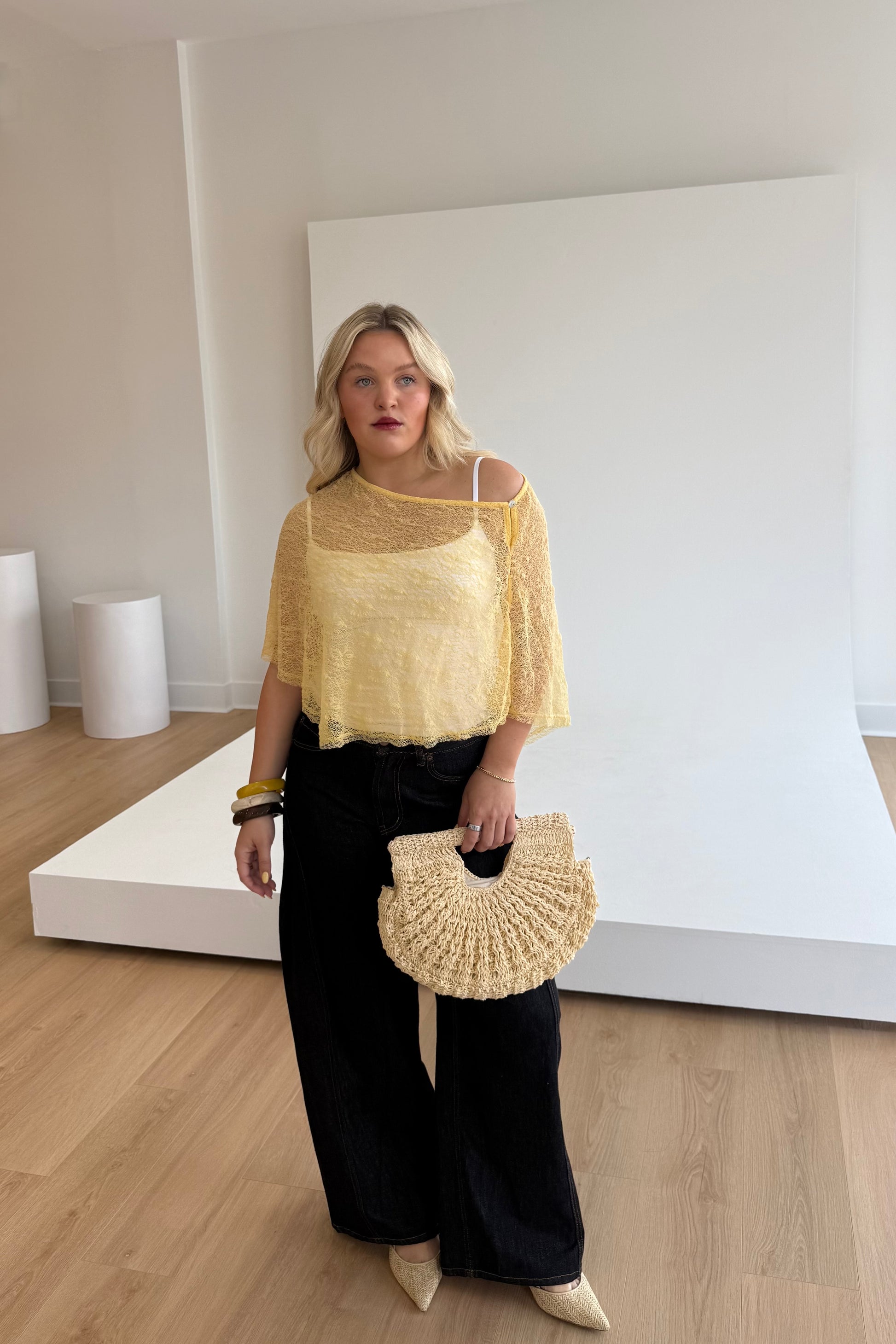 Woman wearing a yellow top and black pants, holding a beige handbag in a minimalistic room.