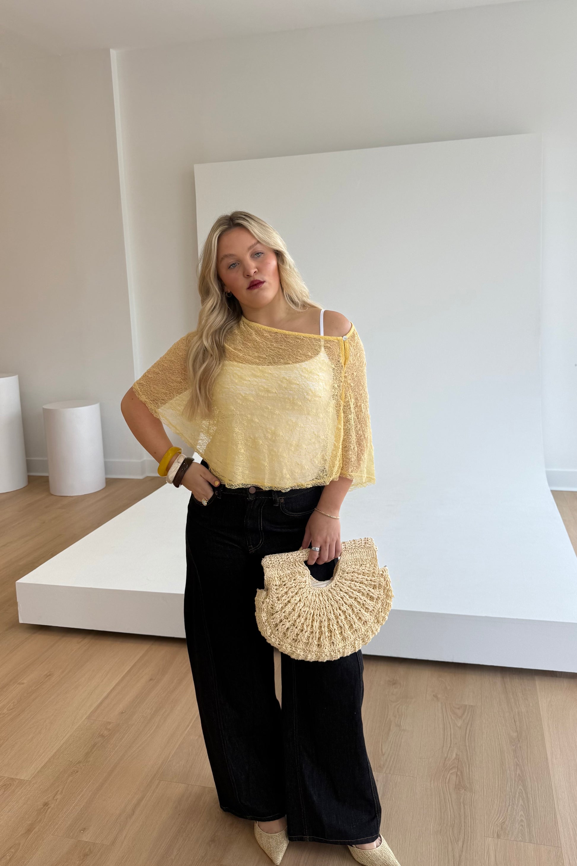 Woman wearing a yellow top and black pants, holding a beige handbag in a minimalistic setting.