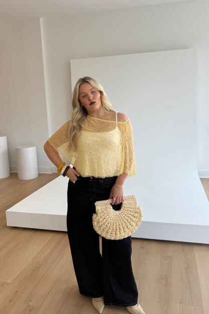 Woman wearing a yellow top and black pants, holding a beige handbag in a minimalistic setting.