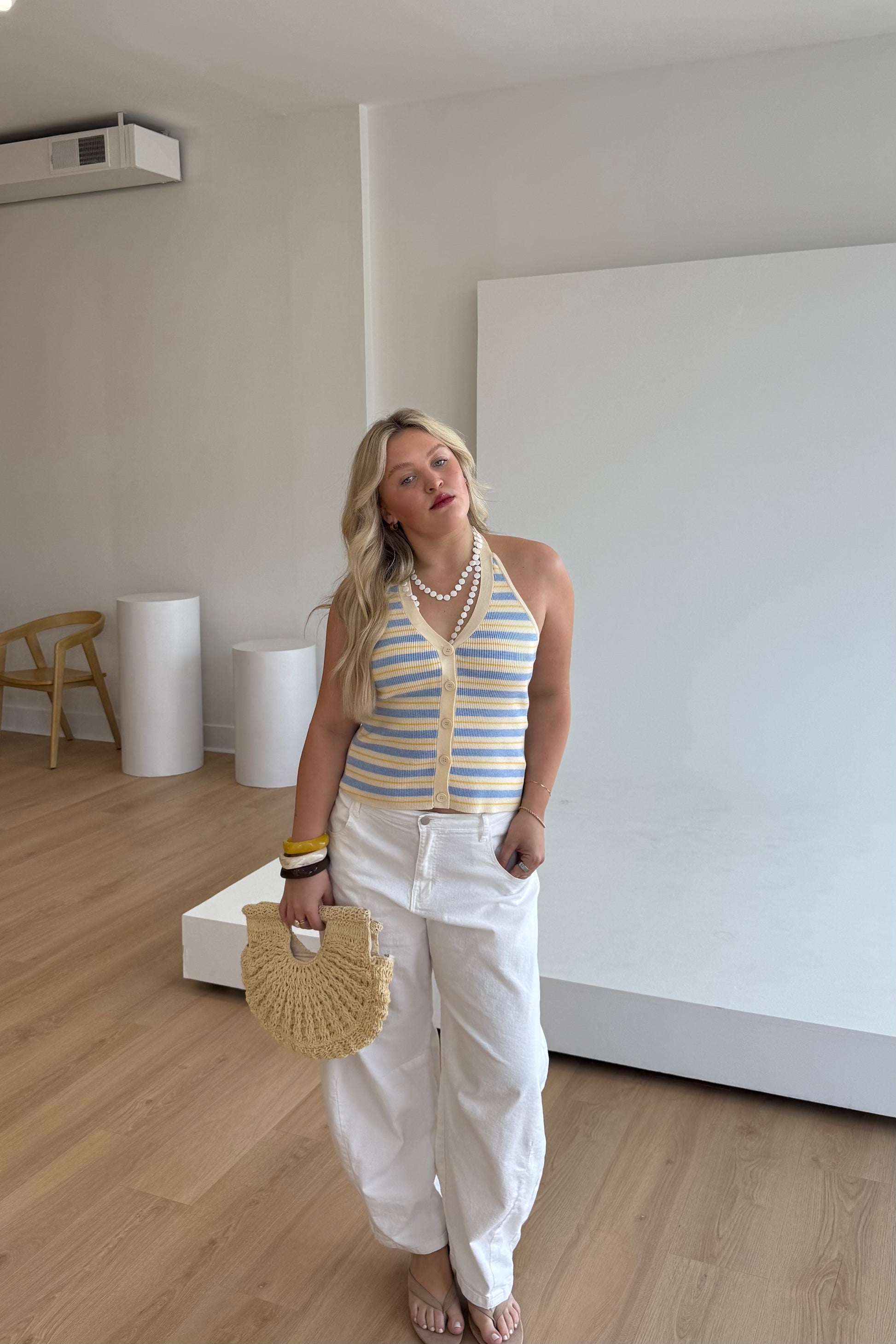 Woman in a striped top and white pants holding a woven handbag in an indoor setting.