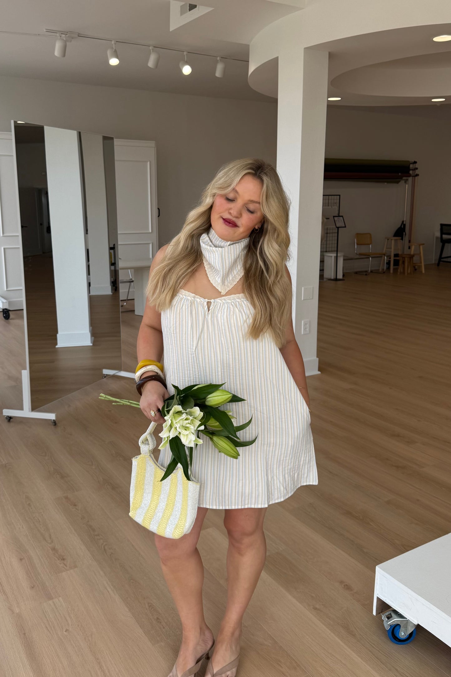 Woman in a white dress holding flowers and a striped bag in an indoor setting.