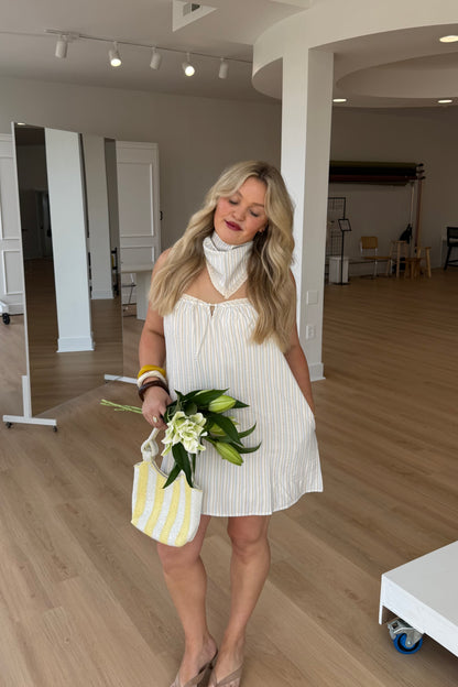 Woman in a white dress holding flowers and a striped bag in an indoor setting.