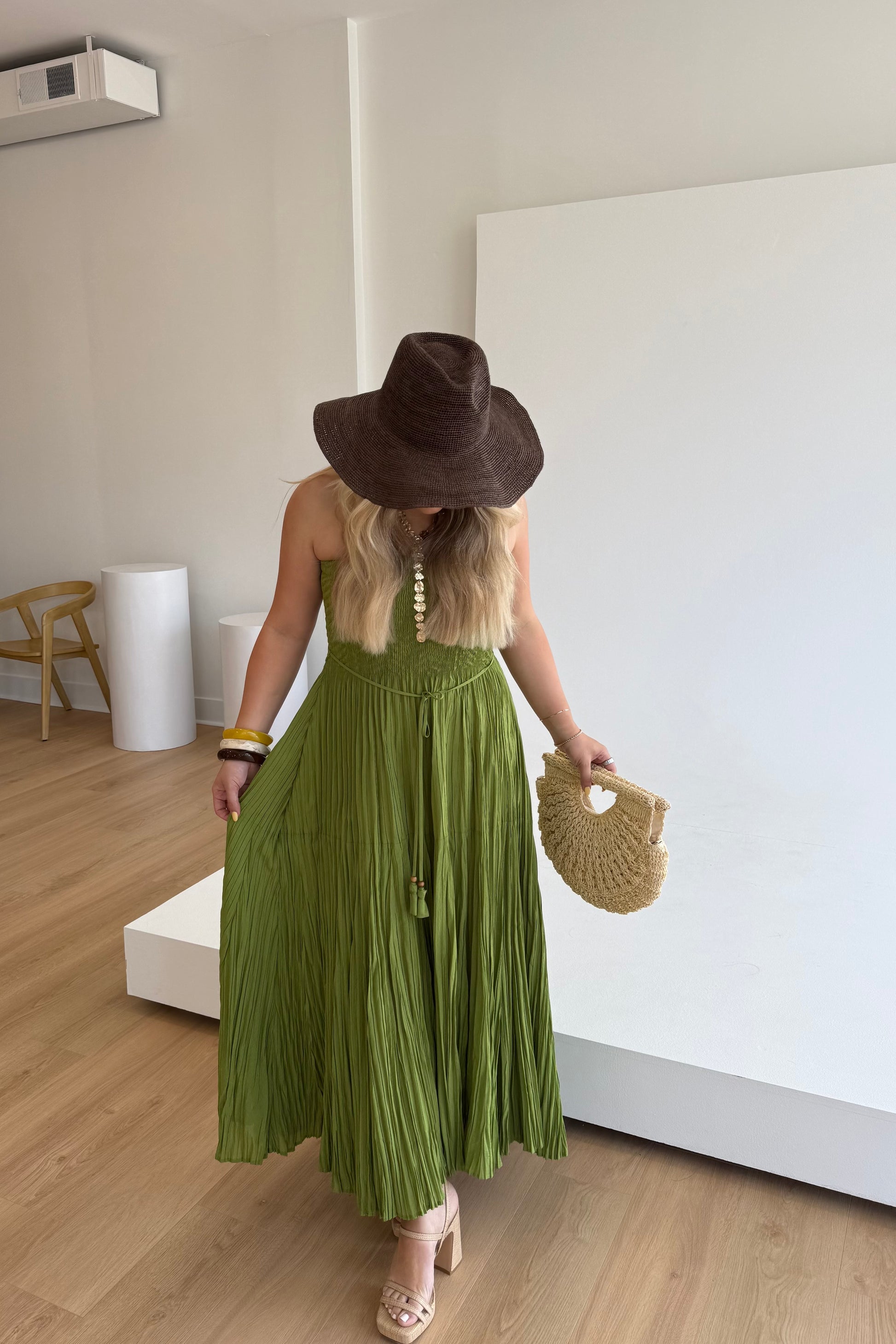 Woman in a green dress and brown hat holding a straw bag in a minimalistic room.