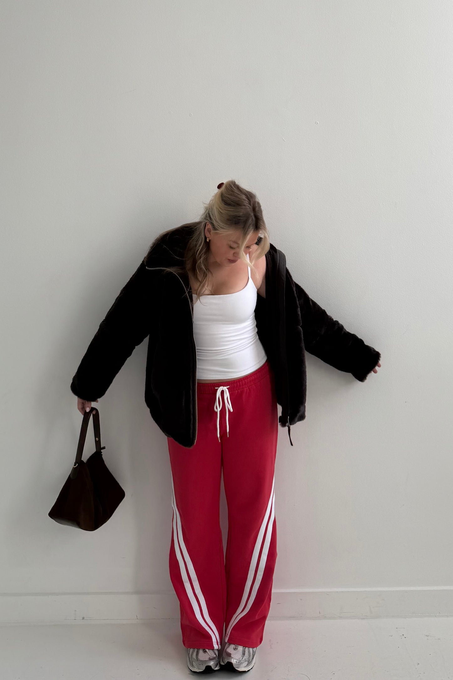 Woman in black fuzzy jacket, white tank top, and red joggers with white stripes, holding a brown bag.