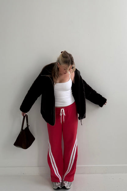 Woman in black fuzzy jacket, white tank top, and red joggers with white stripes, holding a brown bag.