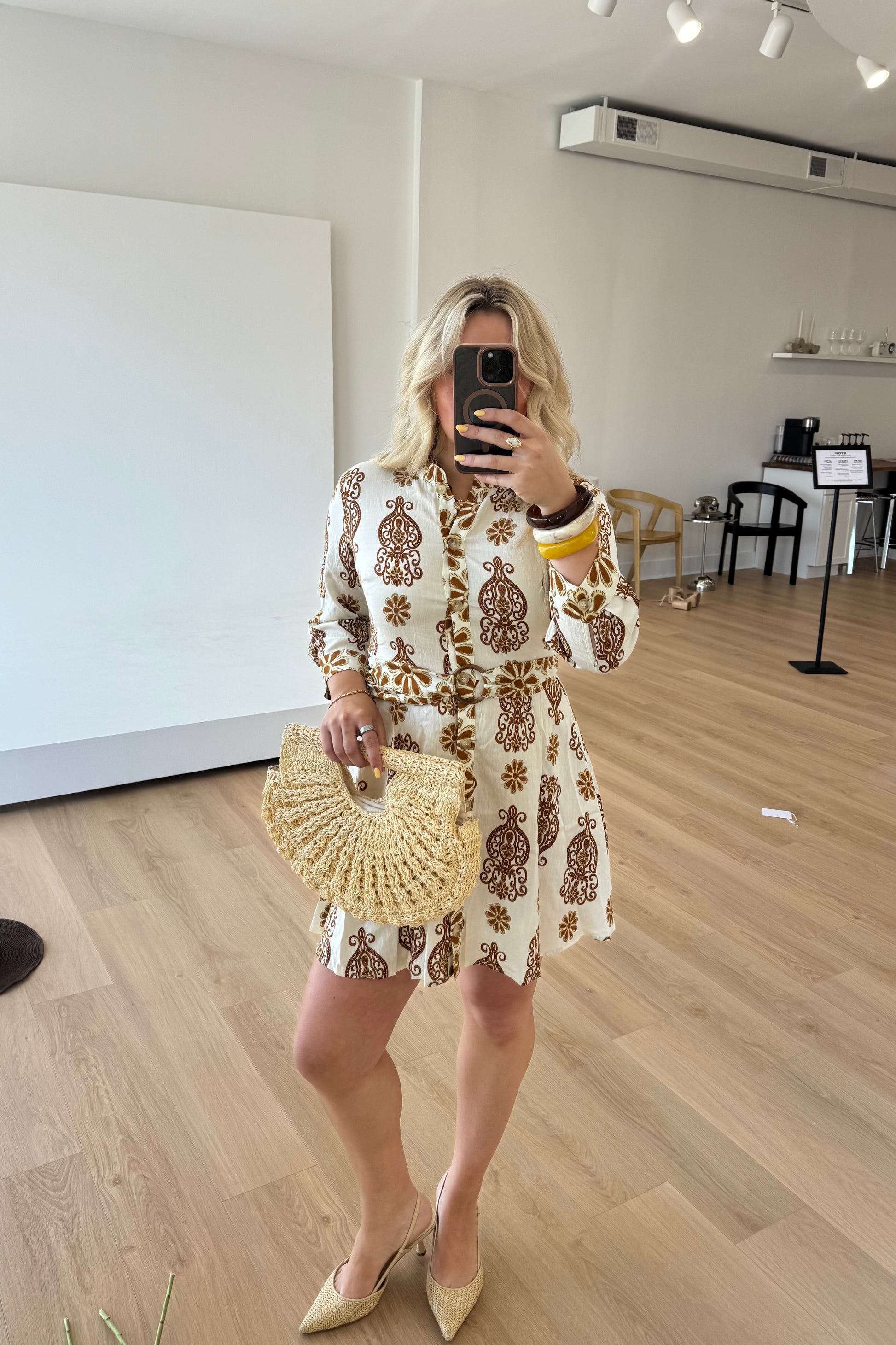 Woman taking a mirror selfie wearing a patterned dress and holding a woven handbag in an indoor setting.