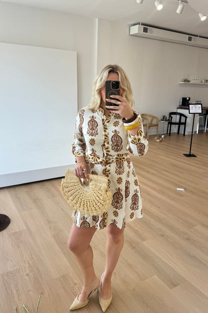 Woman taking a mirror selfie wearing a patterned dress and holding a woven handbag in an indoor setting.