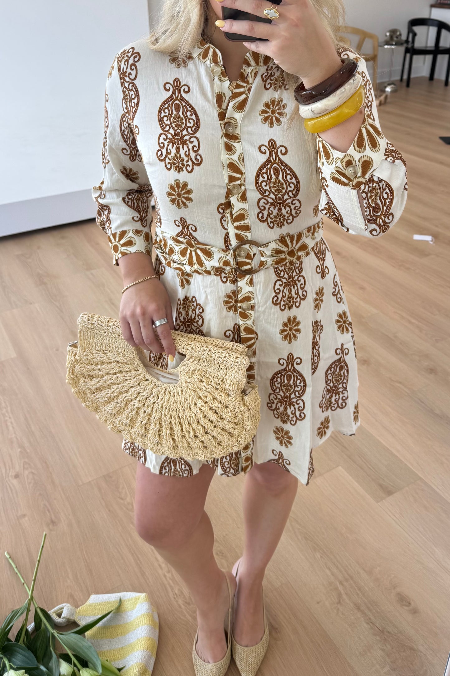 Woman in a white dress with gold embroidery holding a straw clutch on a wooden floor.