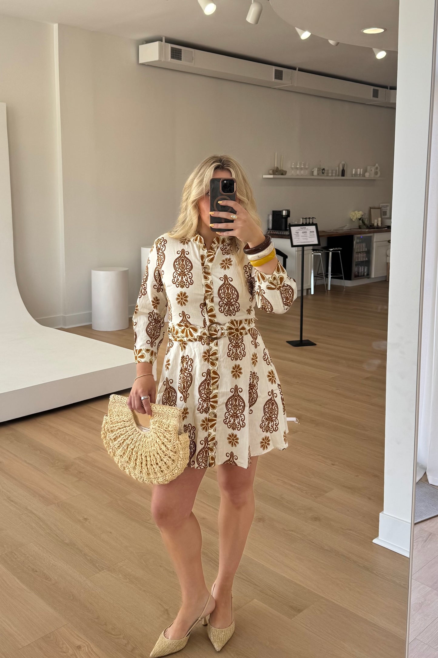 Woman taking a mirror selfie wearing a patterned dress and holding a straw bag in a room with wooden flooring.