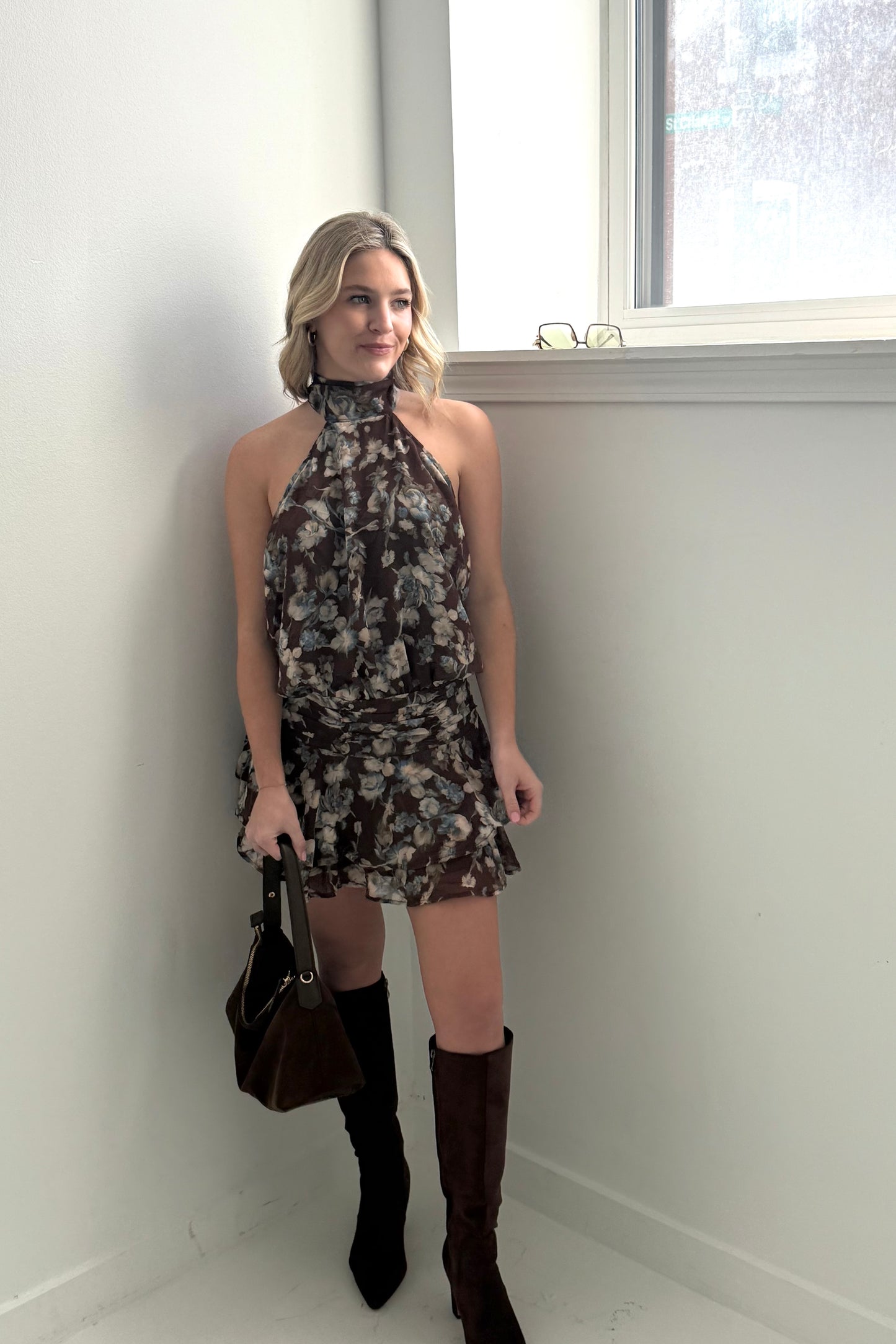 Floral halter neck dress in brown with ruffled skirt, paired with tall black boots and a handbag.