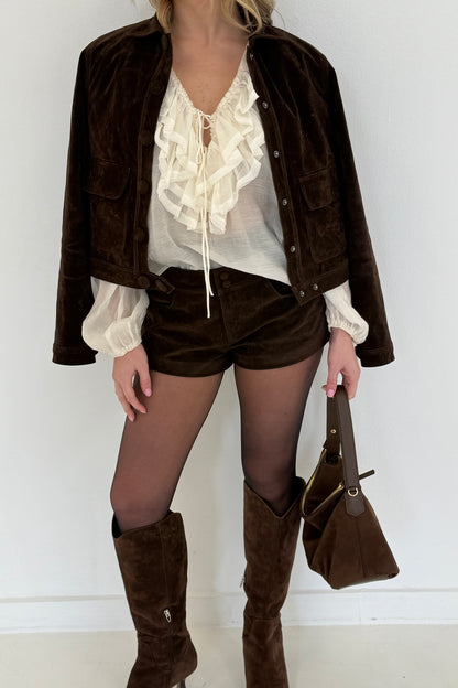 Brown suede jacket, ruffled cream blouse, brown shorts, knee-high boots, and handbag for a chic look.