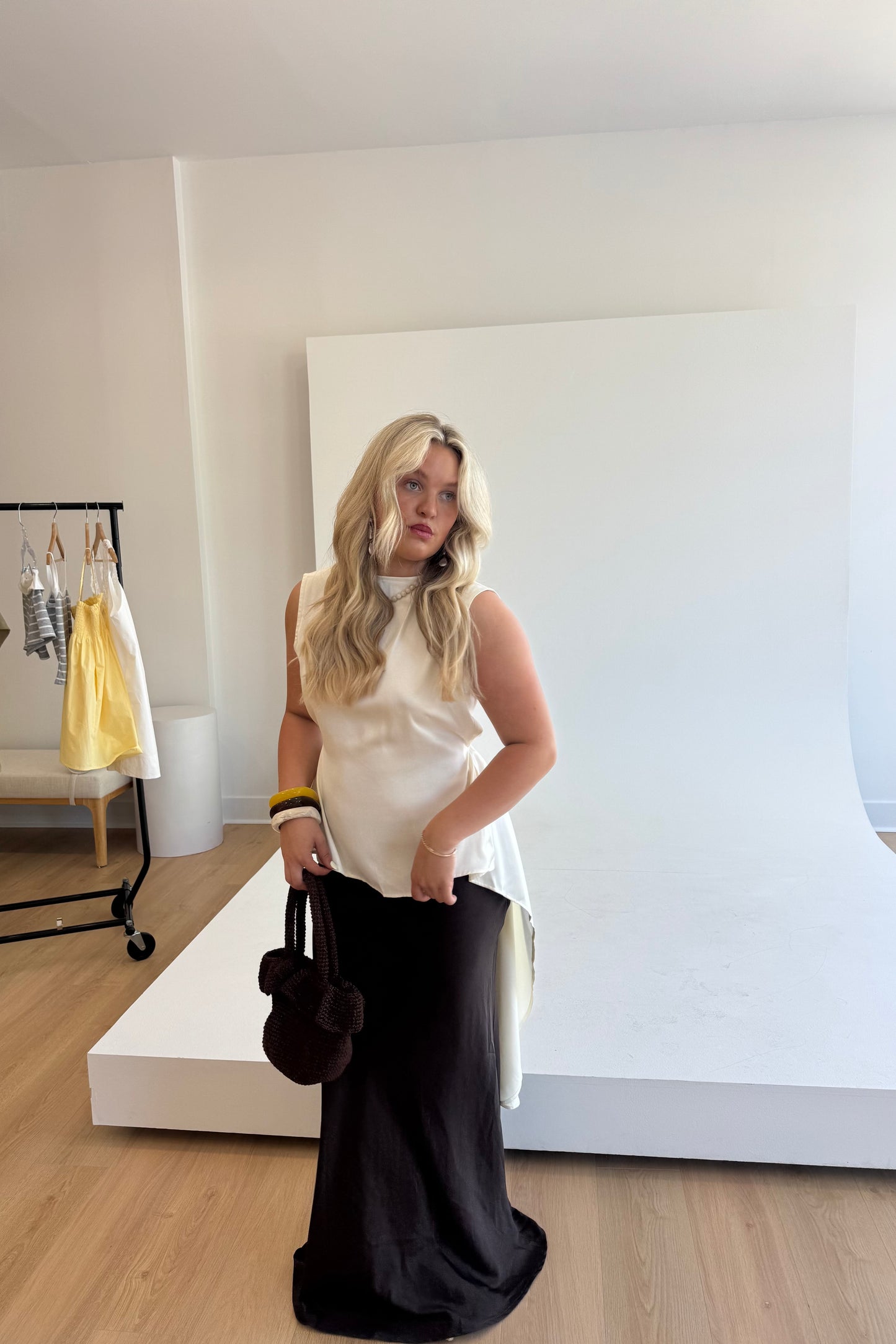 Woman in a white sleeveless top and black pants standing in a room with a white wall and wooden floor.